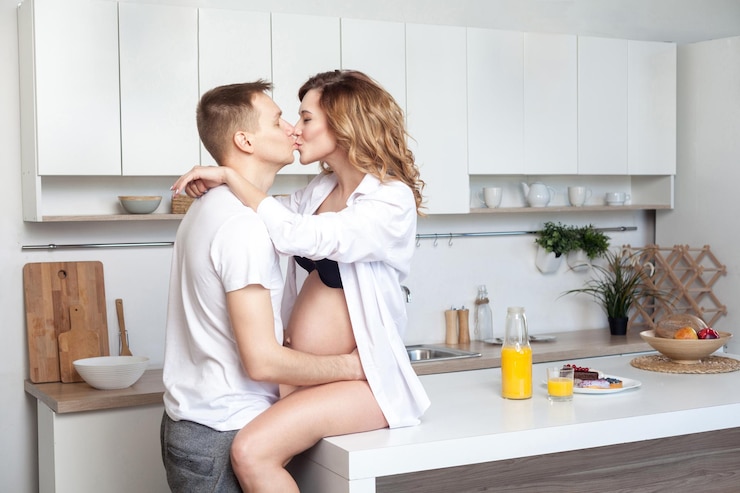 Photo lovestory. young married couple embraces siting on table in kitchen. husband hugs his pregnant wife, putting his hands on her big belly, parents enjoy each other and kissing with closed eyes.