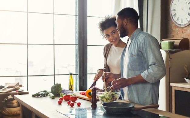 Loving afro american couple preparing green salad for dinner together in loft kitchen