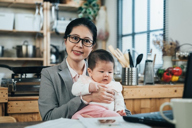 彼女の愛らしい子供を抱きしめる愛情のあるアジアの母親は幸せそうに笑っています。フォーマルなスーツを着た中国人女性が、かわいい赤ちゃんを心地よい表情で抱いている。