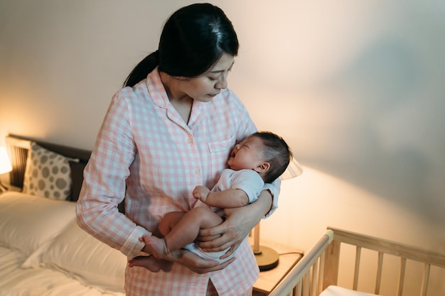 loving asian mother is looking at her upset baby in arms and comforting it with gentle pat by the crib at bedtime in a cozy bedroom at home.