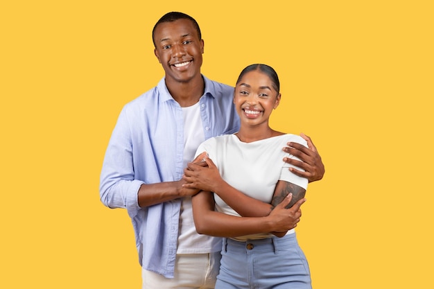 Loving black man hugging his wife from back standing and smiling together over yellow studio