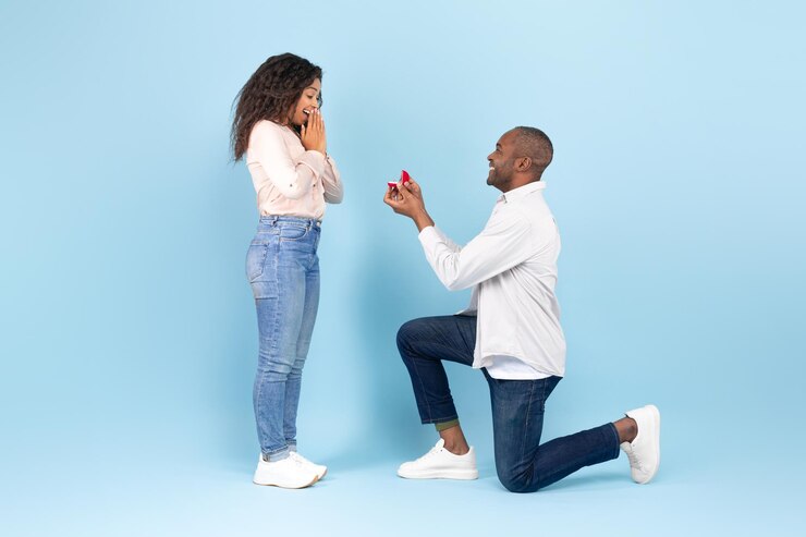 Photo loving black man standing on one knee and offering engagement ring to his beloved woman on blue background side view