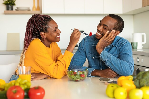 Loving black woman feeding her boyfriend with fresh salad eating in cozy kitchen together having lunch at home