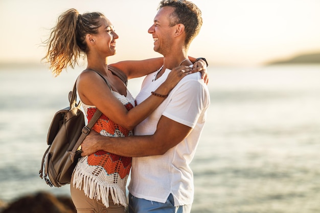 愛情のある陽気なカップルは、夏休みを楽しんで、日没時に空の海のビーチで抱き締めています。