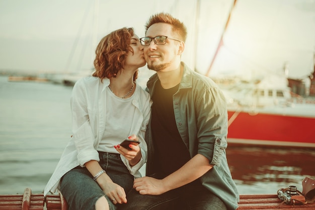 Loving cheerful couple of lovers kiss while sitting on a bench against the background of  yachts club at sea