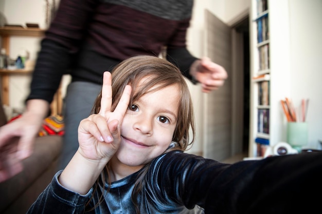 Loving child girl taking selfie by phone with dad indoor in living room making victory sign