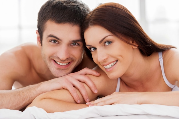 Loving couple in bed. Cheerful young loving couple lying in bed and smiling at camera