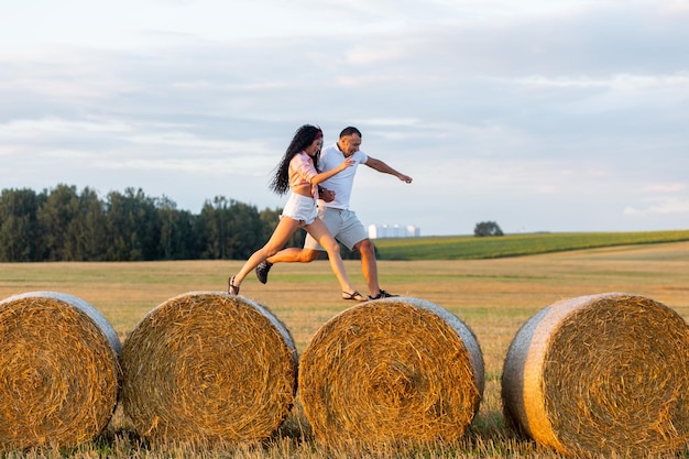Влюбленная пара в поле на рулонах соломы. Молодой мужчина и женщина веселятся, прыгают и позируют на закате.