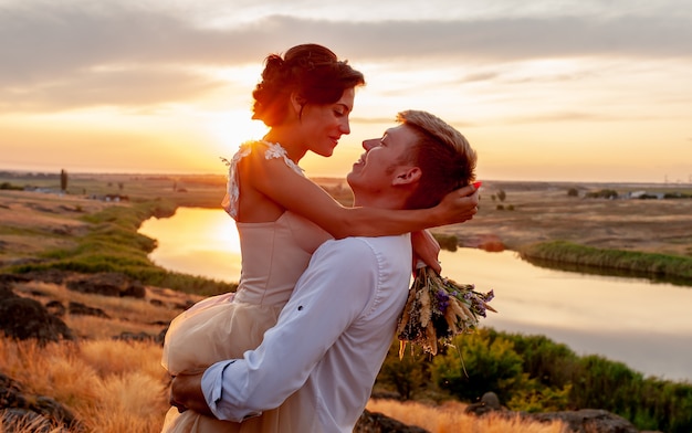 愛するカップル、男と女のキスと川の背景に山の夕暮れ時に抱擁