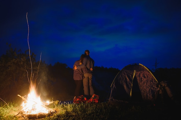 Coppia di innamorati escursionisti che si godono a vicenda, in piedi accanto al fuoco di notte sotto il cielo serale vicino a alberi e tenda