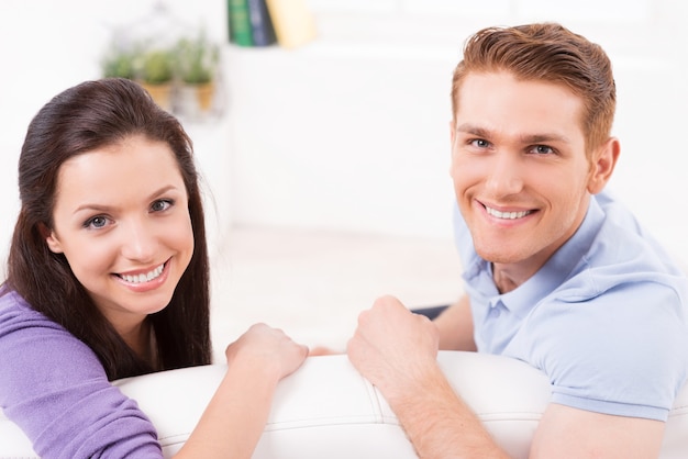 Loving couple at home. Top view of beautiful young loving couple sitting together on the couch and looking at camera with smile