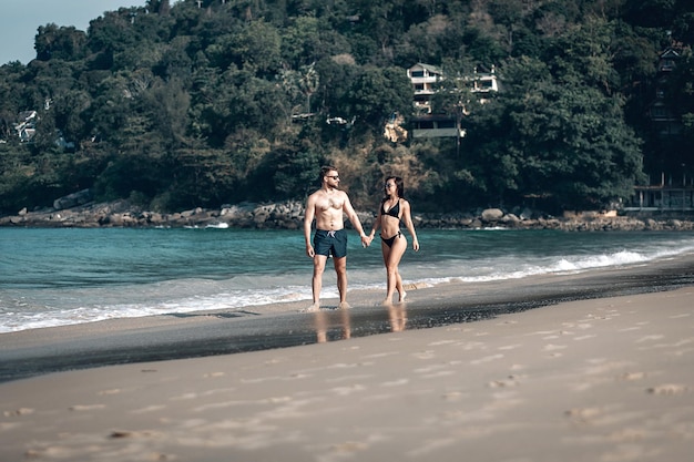 Loving couple on honeymoon walks along the sunny exotic beach holding hands, sunglasses and swimwear on; romance concept.