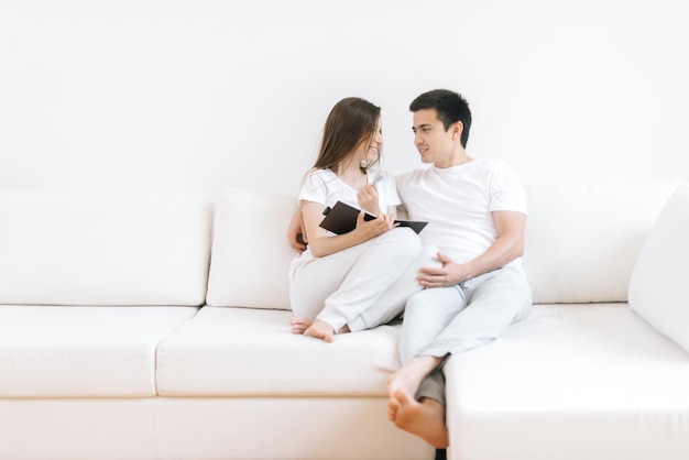 Loving couple hugging holding hands relaxing on cozy sofa. Smiling boyfriend and girlfriend looking at each other embracing at couch. Young happy lovers resting enjoying weekend at home together