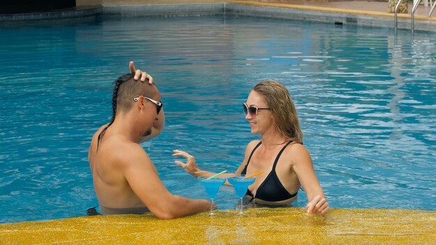 The loving couple hugs and kisses drinking blue cocktail alcohol liquor in swimming pool at hotel outdoor Portrait of caucasian man and woman Creative hairstyles bodybuilder swimsuit sunglasses