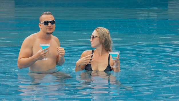 The loving couple hugs and kisses drinking blue cocktail alcohol liquor in swimming pool at hotel outdoor Portrait of caucasian man and woman Creative hairstyles bodybuilder swimsuit sunglasses