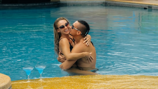 The loving couple hugs and kisses, drinking blue cocktail alcohol liquor in swimming pool at hotel outdoor. Portrait of caucasian man and woman.