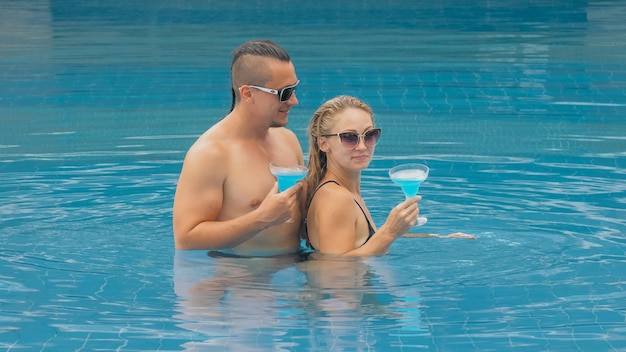 The loving couple hugs and kisses drinking blue cocktail alcohol liquor in swimming pool