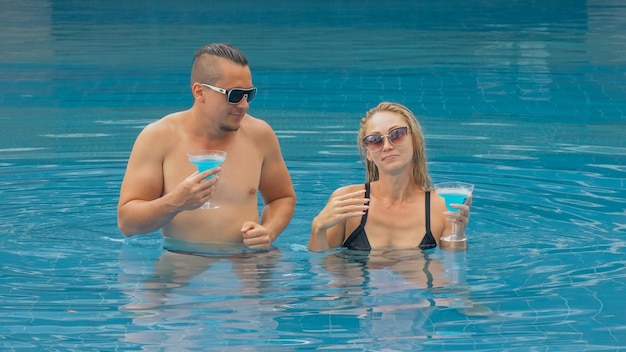 The loving couple hugs and kisses drinking blue cocktail alcohol liquor in swimming pool