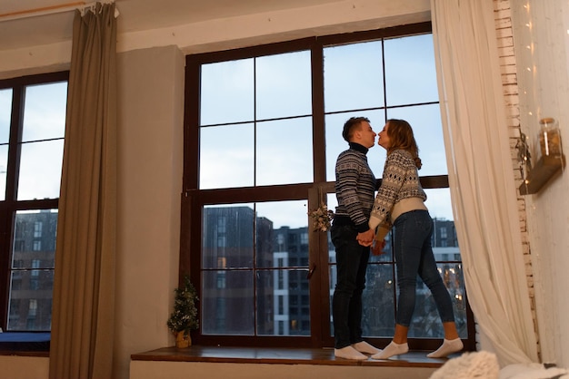 A loving couple in knitted sweaters stand by the hand against the background of a large window on the windowsill