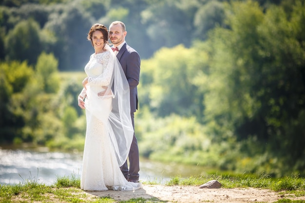 Loving couple of newlyweds walks near wide river in summer sunny day