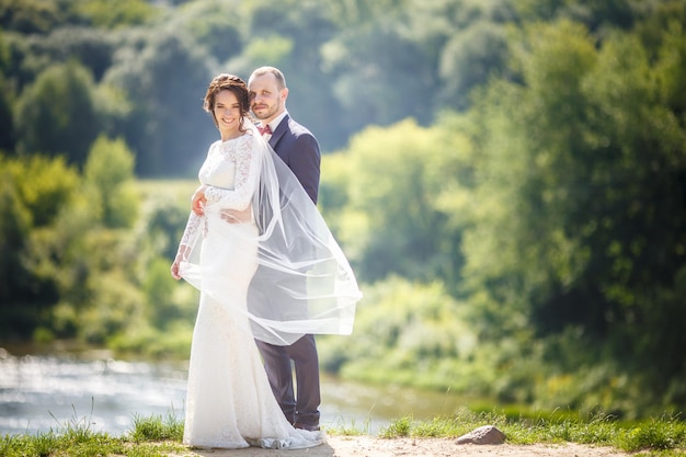 Loving couple of newlyweds walks near wide river in summer sunny day