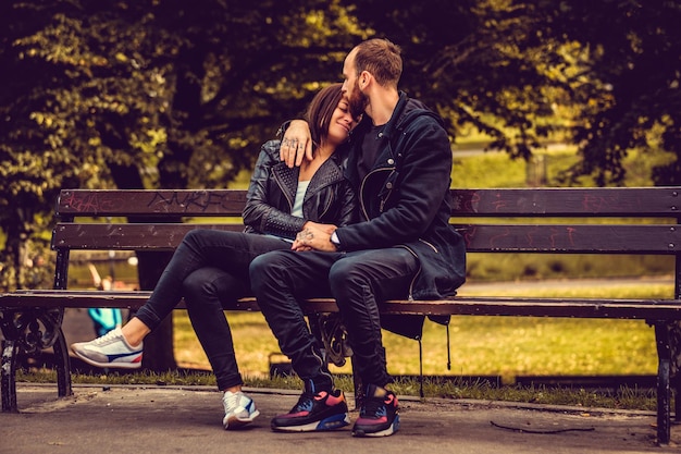 Loving couple posing on a bench in autumn city park. Bearded male hugging attractive brunette female.