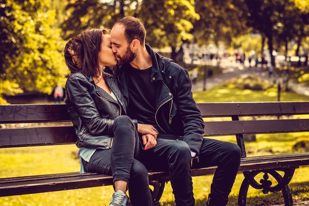 秋の公園のベンチでリラックスした愛情のあるカップル。若いブルネットの女性にキスするカジュアルなひげを生やした男性。