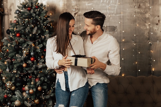 Coppia di innamorati in piedi nel soggiorno accanto a un albero di natale splendidamente decorato con ghirlanda. la donna ha ricevuto un regalo dall'uomo.