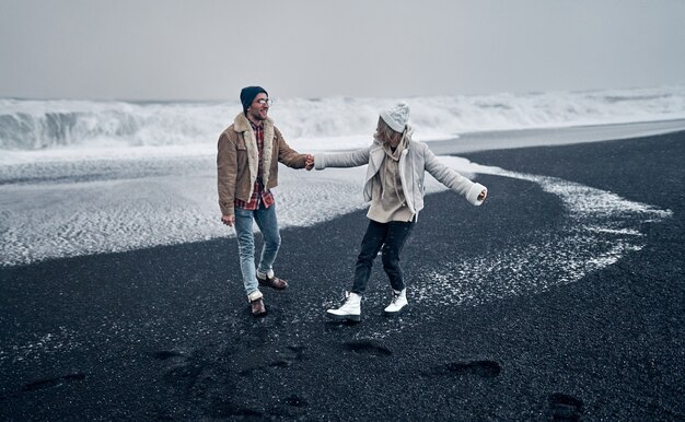 A loving couple of tourists holding hands have fun while\
walking along the beach with black volcanic sand and admire the\
great ocean waves. travel, leisure, tourism.