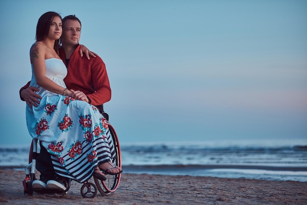 A loving couple, a woman sits on her husband's lap, resting on a beach against a background of a bright dawn.