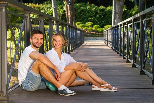 木製の橋の上の愛情のあるカップルと橋の上に座っている女性の若いカップルは、楽しい時間を過ごしています...