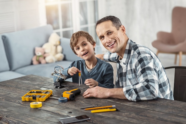 Loving daddy. Attractive cheerful dark-haired man showing instruments to his son while sitting at the table and his son holding compasses and smiling