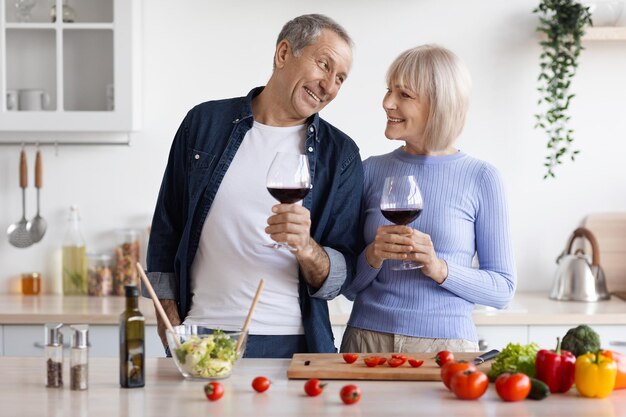 Photo loving elderly spouses drinking wine while cooking together