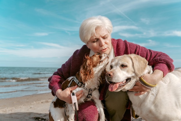 Loving gray-haired Caucasian senior woman hugging her two adorable canine friends on the sandy beach