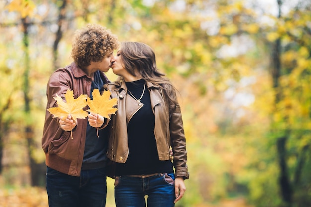 秋のカエデの葉を手に持って公園で秋の幸せなカップルを愛する。男と女のキス