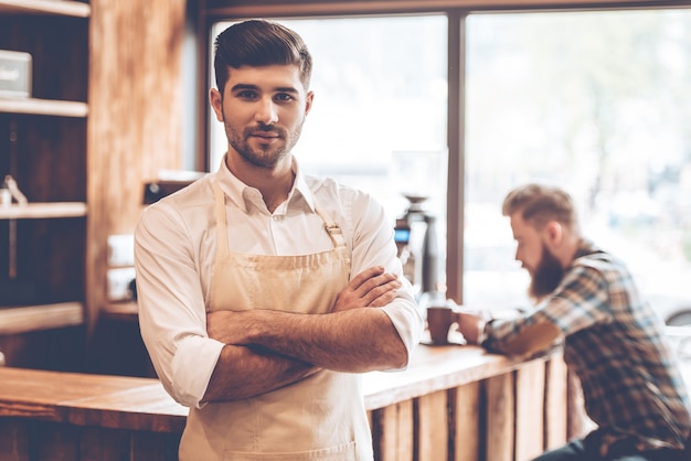 Loving his work. Young handsome man in apron looking at camera and keeping arms crossed while standing at cafe with customer at background