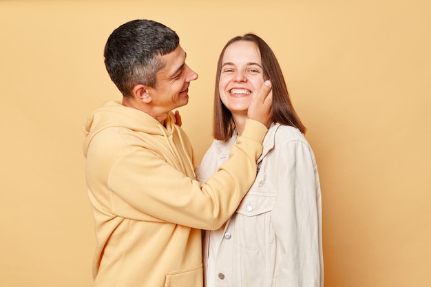 Loving husband touching wife face looking with love at his lovely pretty girlfriend woman and man wearing casual style clothing standing isolated over beige background