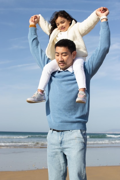 喜欢拍照的日本家庭在海滩上散步。女儿骑在父亲的背上，望着前方。休闲、度假、育儿理念