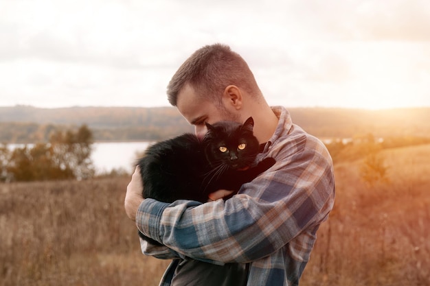 秋の森で愛する男と彼の黒い猫 彼は自分のペットを肩に背負って公園でリードで歩いています