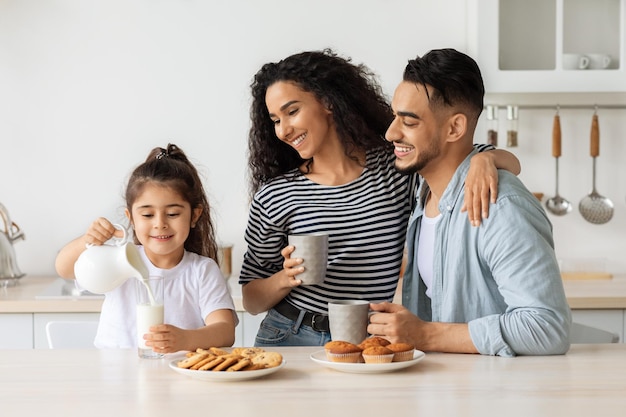 Loving middle-eastern family of three enjoying healthy breakfast together, beautiful curly mother hugging her handsome husband and looking at pretty little daughter pouring milk into glass, copy space