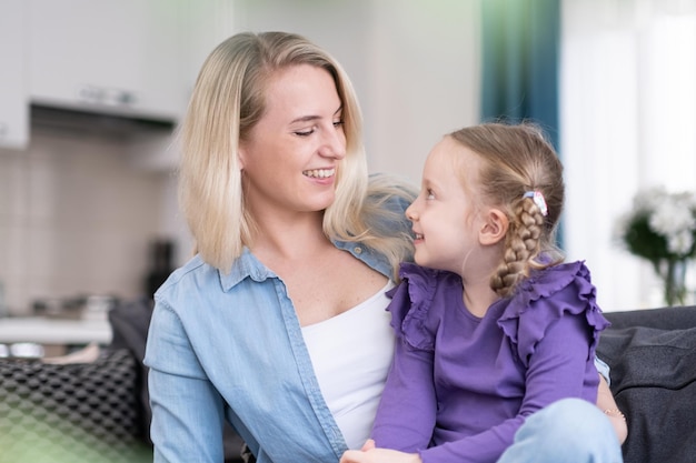 Loving mom with cute little daughter sitting on sofa at home hugging and smiling happy family