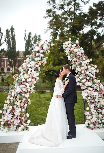 Влюбленные молодожены стоят возле цветочной аркиМолодая красивая счастливая свадебная пара целуется на церемонии