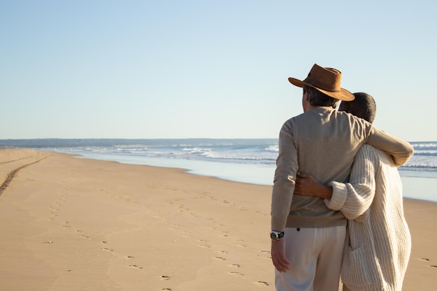 Loving senior couple standing at seashore, hugging each other and enjoying seascape view. Grey-haired man in cowboy hat embracing his wife while spending holiday together. Love, retirement concept