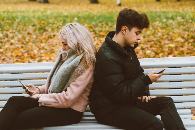 Loving teenagers on date look at mobile phones, sit on park bench in fall.