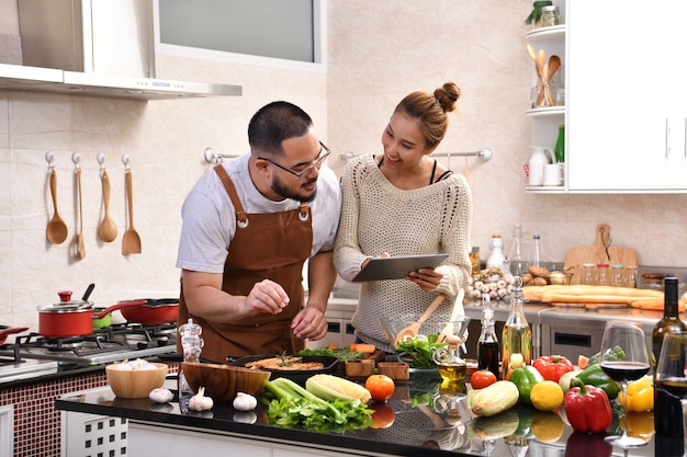 Loving young Asian couple using digital tablet and cooking in kitchen making healthy food together feeling fun