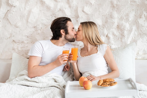 Loving young couple having breakfast in bed together kissing spending honeymoon at hotel