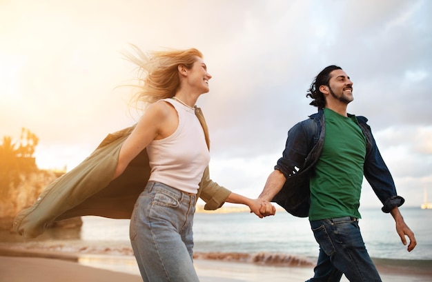 Loving young european couple holding hands while running on beach at sunset