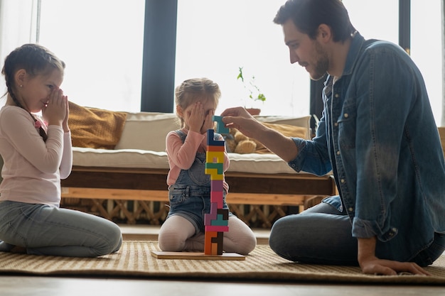 Loving young father lying on floor with small kids have fun engaged in funny activity with construction bricks, caring dad play with little children build with blocks enjoy time at home together