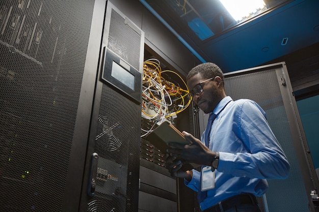 Low angle of African American man holding digital tablet while standing by server cabinet and working with supercomputer in data center, copy space