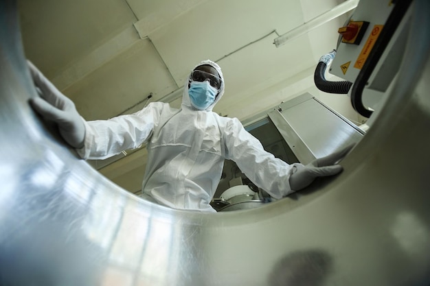 Low angle of black man wearing protective suit looking in tank at factory